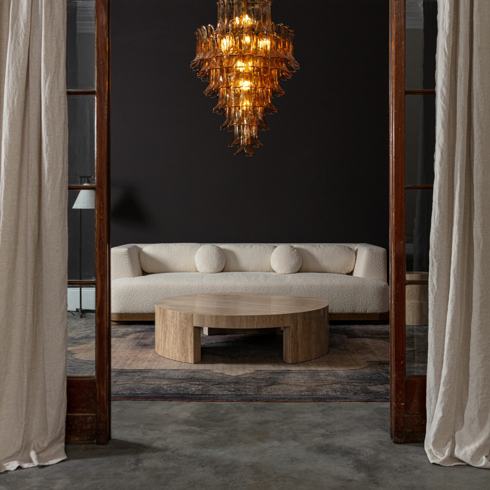 Amber Corolla Large Chandelier Modern living room featuring a plush cream sofa, circular wooden coffee table, and a stunning glass chandelier. Maison Mermont.